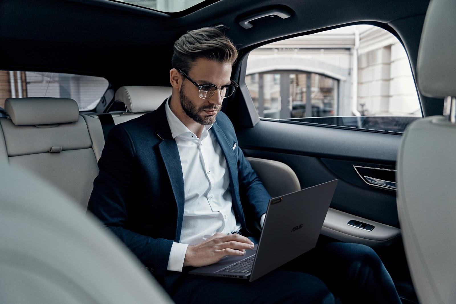 A person in a suit and glasses sitting in a car using a computerDescription automatically generated
