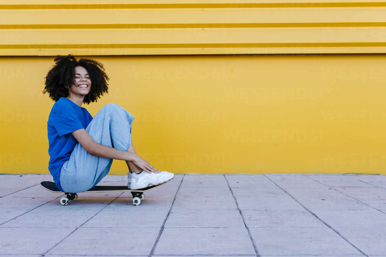 Smiling young woman sitting on skateboard in front of yellow wall stock photo